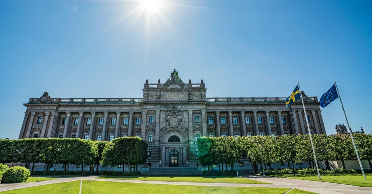 Front view of the Riksdag building with Swedish and EU flags on a sunny day.